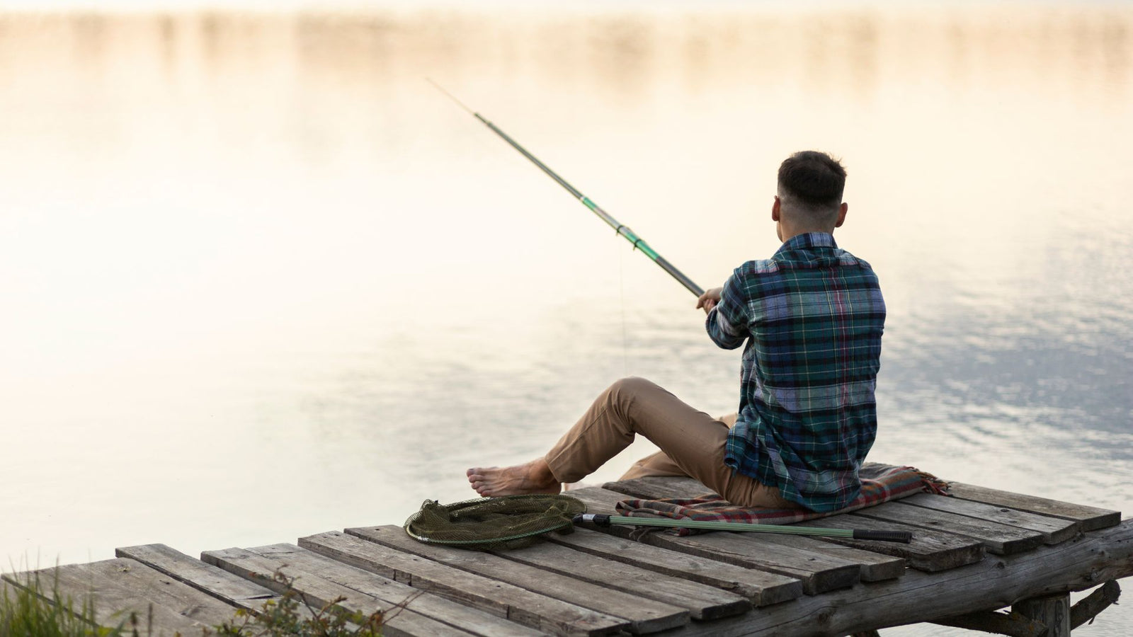  Man fishing by the river. He is sitting on a small dock and holding a fishing rod with his hand, while looking at the water. The background features trees and a calm river.